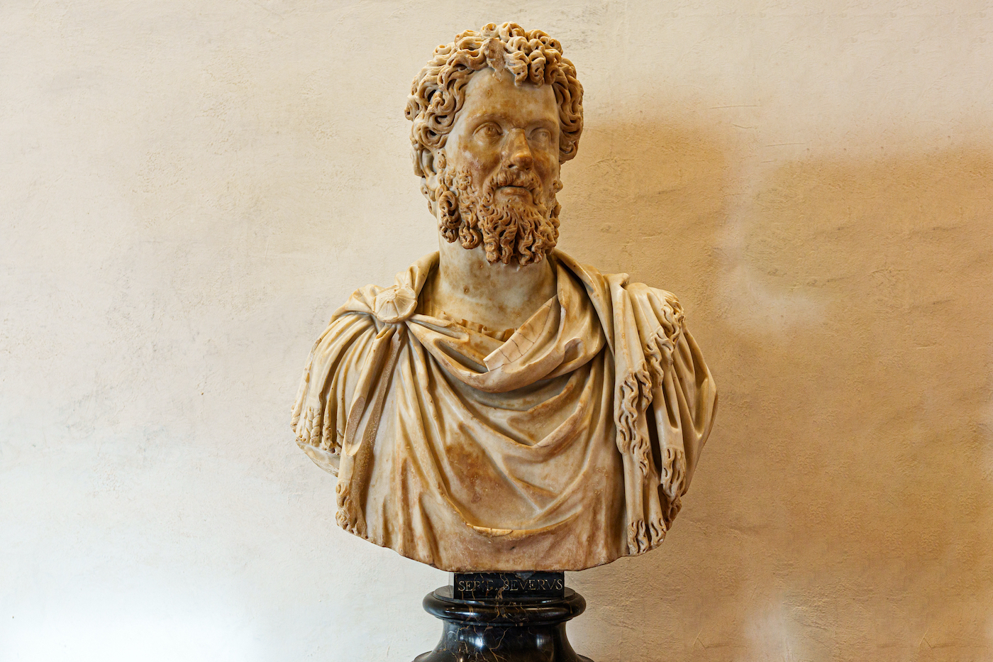 Busto de mármol de Septimio Severo, emperador romano del siglo III, con barba y cabello ondulado, vistiendo paludamentum (manto militar) sobre una armadura, montado sobre un pedestal negro de mármol