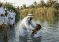 Recreación de un bautismo mandeo en un río del sur de Irak: un sacerdote con barba blanca, turbante y corona de mirto sumerge a un fiel vestido de blanco, mientras hombres, mujeres y niños con túnicas blancas y coronas de mirto esperan en la orilla junto a una mesa de caña con pan.