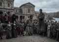Recreación de la ejecución de Prisciliano en Tréveris: un hombre con túnica y las manos atadas, arrodillado ante un verdugo con espada, frente a una multitud y soldados romanos, mientras el emperador con manto púrpura y varios obispos observan desde una tribuna junto a un río.